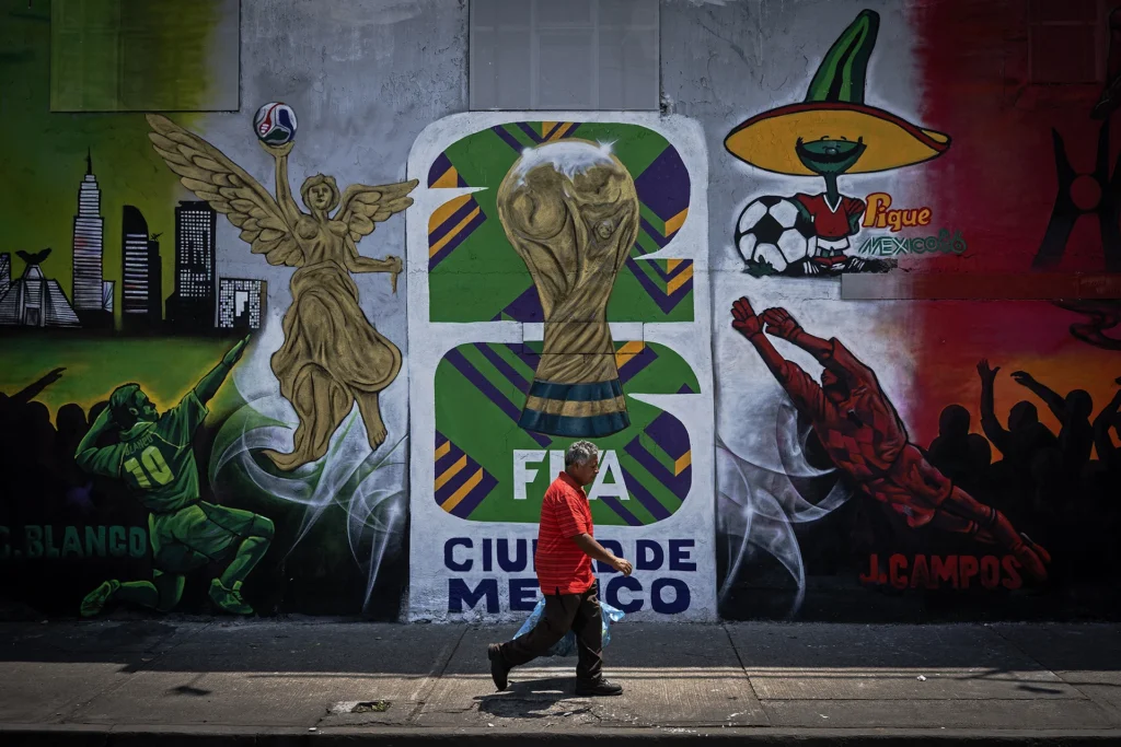 FIFA World Cup 2026: The Geopolitical Tensions at Play Off the Pitch A man walks along a sidewalk past a vibrant outdoor street mural in Mexico City depicting traditional soccer imagery next to the official tournament logo, showcasing the local sports culture as geopolitical tensions and conflict impacts loom over the FIFA World Cup.