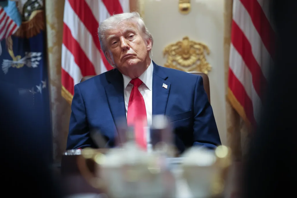 مسيرة الحماقة تصل إلى هرمز President Donald Trump sits at a conference table inside the brightly lit White House Cabinet Room flanked by American flags, his expression pensive as geopolitical strategies take shape concerning international maritime chokepoints like Hormuz.