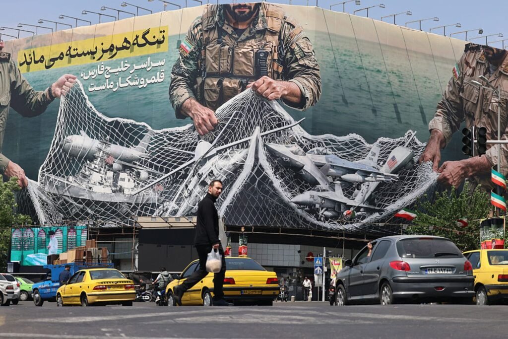 Was It Worth It? The True Cost of Trump’s Iran War Vehicles drive past a giant propaganda billboard at Revolution Square in Tehran depicting a soldier capturing military assets in a net, visually representing the domestic mobilization and geopolitical fallout tied to the True Cost of Trump's Iran War.