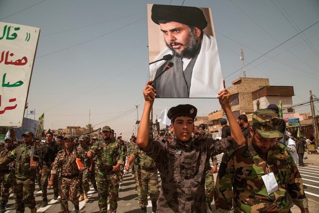 Sadr once again dismantles his armed militia. Why now? A young militia member in a grey camouflage uniform holds a large portrait of Muqtada al-Sadr above his head during an outdoor military parade in Sadr City, Baghdad, leading a column of armed men in camouflage attire, illustrating the factional dynamics evaluated when Sadr dismantles his militia.