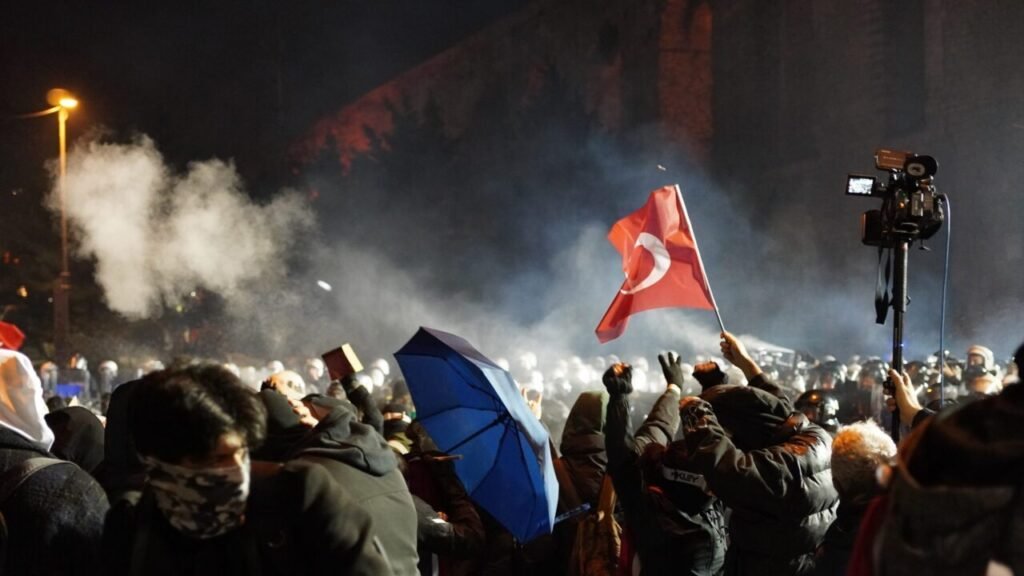 A crowd of young protesters stands facing a line of riot police at night, with one individual waving a Turkish flag and another holding a blue umbrella amidst smoke, illustrating growing public discontent with the Erdogan regime.