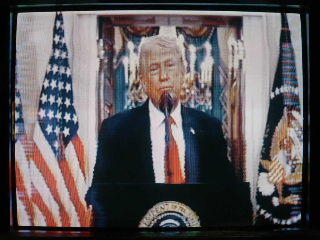 ترامپ حق داشت. جنگ با تسلیم به پایان رسید A television screen displays a broadcast of US President Donald Trump delivering a formal address from a presidential podium flanked by American flags, capturing the televised declaration of how the war ended.