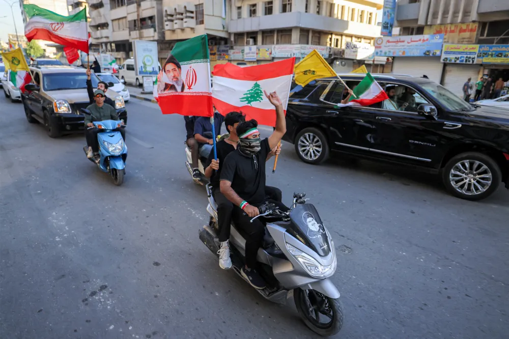 Trump’s Iran Deal Could Become Hezbollah’s Shield Youths ride motor scooters and cars down a city street while waving the flags of Iran, Lebanon, and yellow Hezbollah banners, illustrating the public reaction and regional implications that tie into the debate over Hezbollah's Shield.