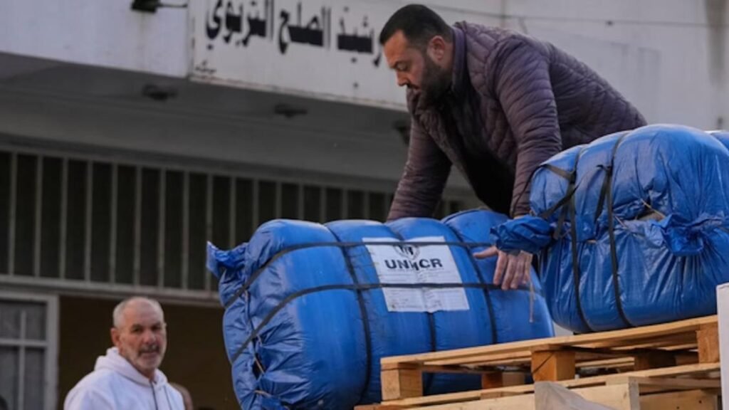 A man in a dark jacket unloading large blue aid bundles labeled with the UNHCR logo from a wooden pallet, highlighting the humanitarian assistance framework supporting refugee populations amidst global aid cuts.