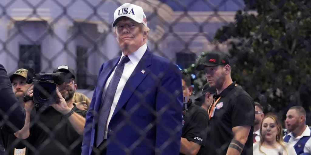 Donald Trump wears a white "USA" hat and dark suit standing behind a chain-link cage fence at the UFC Freedom 250 event on the White House South Lawn after a title bout, symbolizing political optics amid foreign policy evaluations and Trump's failure.