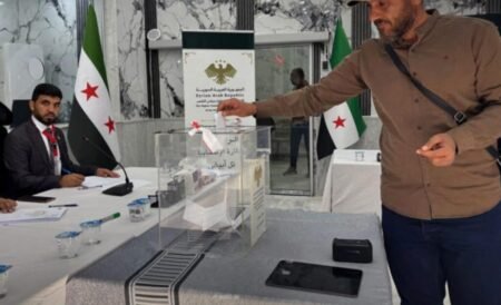A voter casting his ballot into a transparent ballot box at a polling station featuring Syrian national flags and official signs, capturing the voting process as Syria holds elections.