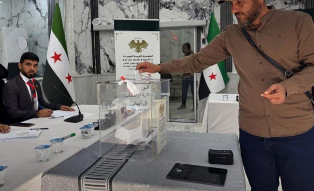 Syria holds elections in Kurdish-majority areas, finds Assad-era chemical weapons A voter casting his ballot into a transparent ballot box at a polling station featuring Syrian national flags and official signs, capturing the voting process as Syria holds elections.