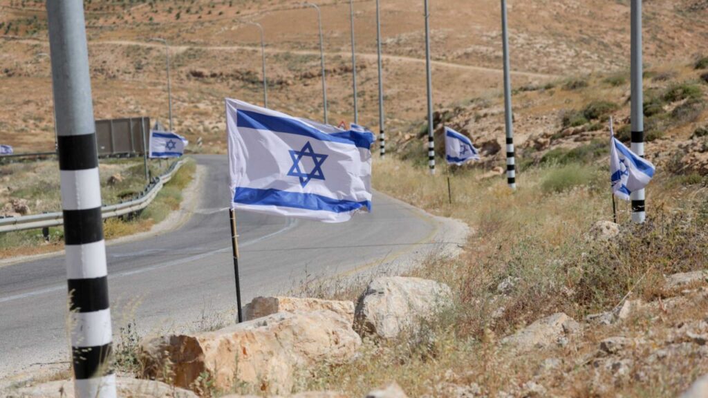 Several Israeli flags fly along the side of a winding asphalt road cutting through an arid, rocky desert landscape, framing geopolitical discussions regarding international legal moves to challenge Israel and its settlements.