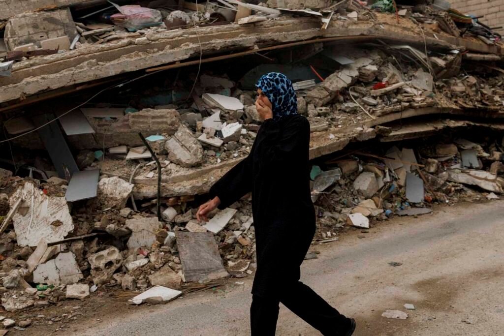 A resident wearing a blue headscarf walking past the heavy concrete rubble of a destroyed building in Zrarieh, Lebanon, covering her nose to protect against the odor rising from the strike site, highlighting the fragile stability under the Israel-Lebanon ceasefire.