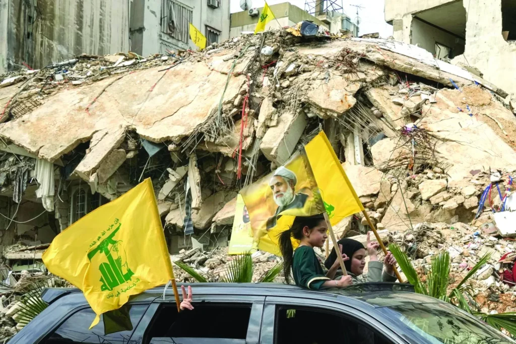 Two young girls wave yellow Hezbollah flags from the sunroof of a car in front of a heavily destroyed concrete building, flashing victory signs amid ruins to underscore the complex socio-political reality behind the mandate to disarm Hezbollah.