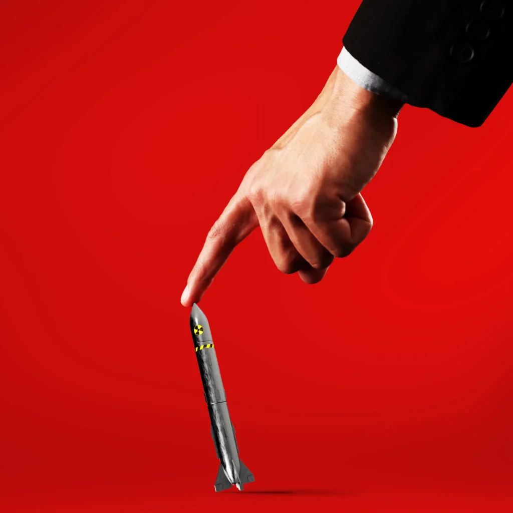این یک توافق بزرگ با ایران نیست. خوب است. A large hand in a business suit points down to touch the tip of a small model missile featuring a nuclear symbol against a solid red background, conceptually representing the limits and intense scrutiny surrounding any potential Grand Bargain With Iran.