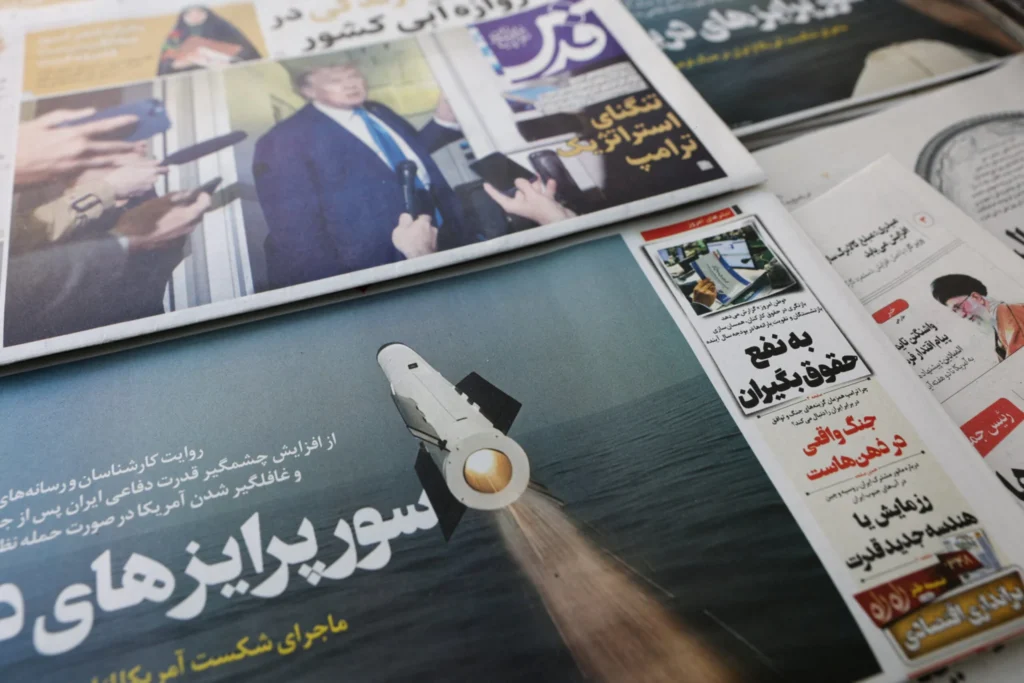 A collection of Iranian newspapers displayed on a surface feature front-page headlines and images covering missile systems and political updates, capturing the shifting regional discourse that follows the announcement of an interim peace deal.