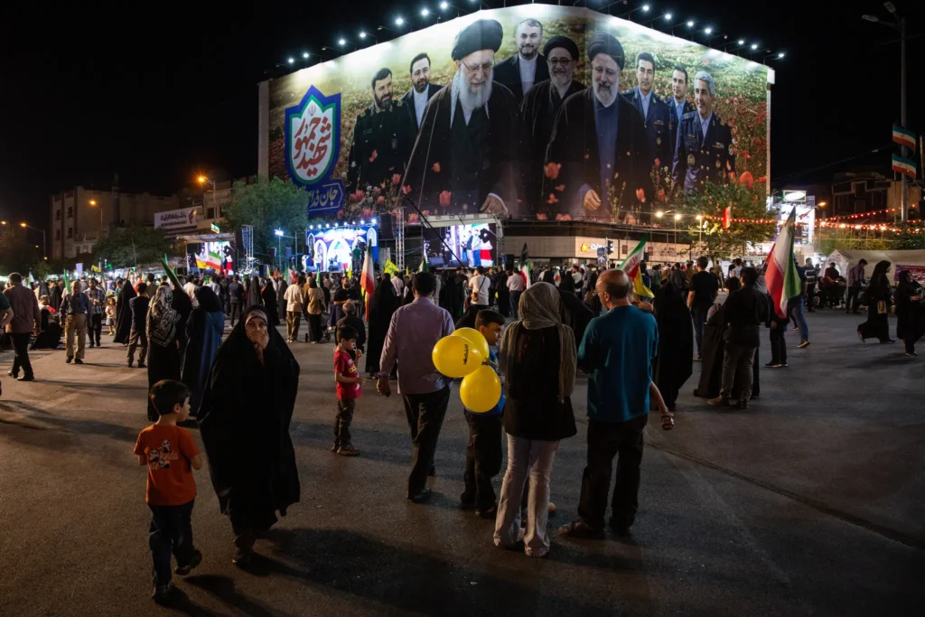A large illuminated billboard in Tehran at night depicting late Iranian leaders, with crowds gathered in the square below as political dynamics shift and the Trump-Led Right Rethinks Iran.