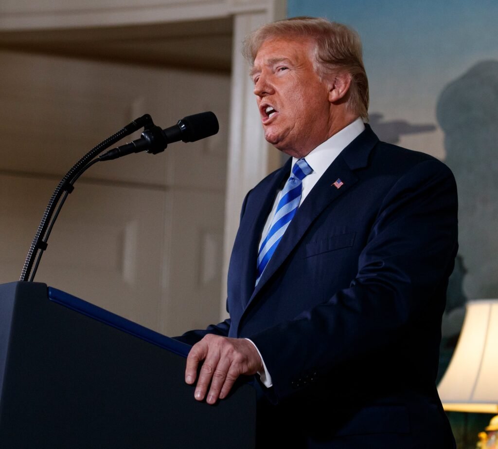 US President Donald Trump wearing a dark suit and striped blue tie speaking at a podium with a microphone, addressing foreign policy challenges linked to Trump's Chaos on Iran.
