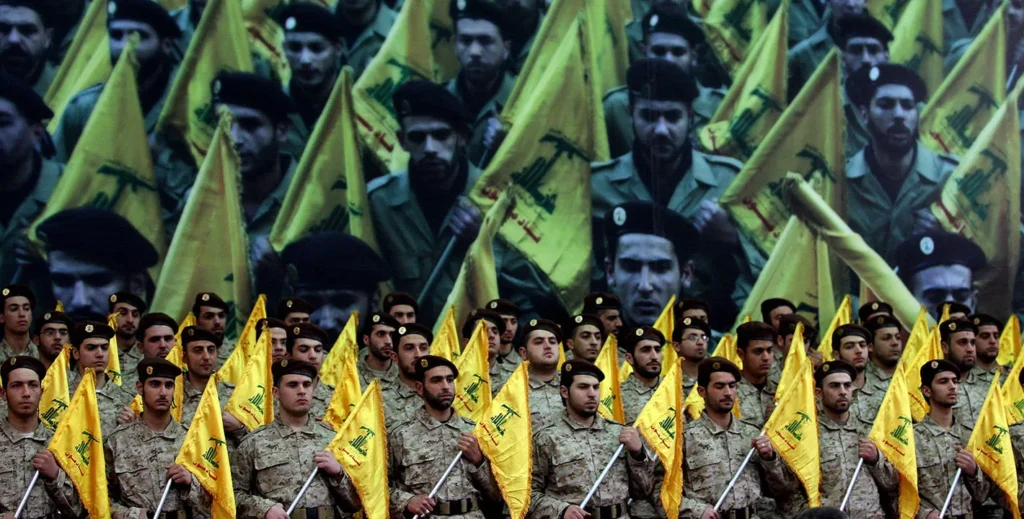 A large crowd of soldiers in military camouflage and black berets standing in rows holding yellow Hezbollah flags, with a massive poster of marching fighters in the background, illustrating regional non-state military presence linked to the analysis of New Syria, Old Lebano