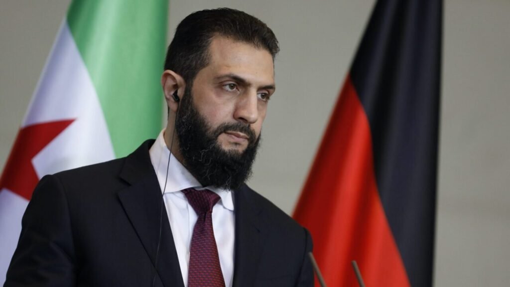 كيف يمكن لسوريا المساعدة في نزع سلاح حزب الله A close-up portrait of Syrian President Ahmed al-Sharaa in a suit and tie with an earpiece, standing between the Syrian opposition and German flags during a press briefing, outlining how Syria Can Help in regional diplomatic shifts.
