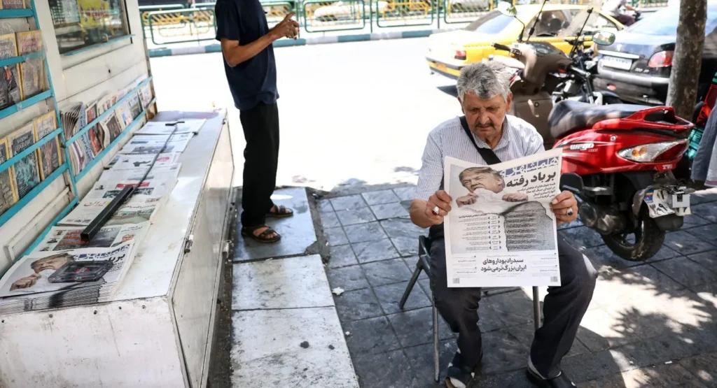 An elderly man sits on a sidewalk chair in Tehran reading a newspaper next to a newsstand kiosk and a parked red motorcycle, capturing the daily domestic perspective during a period when Iran Wanted to Survive.