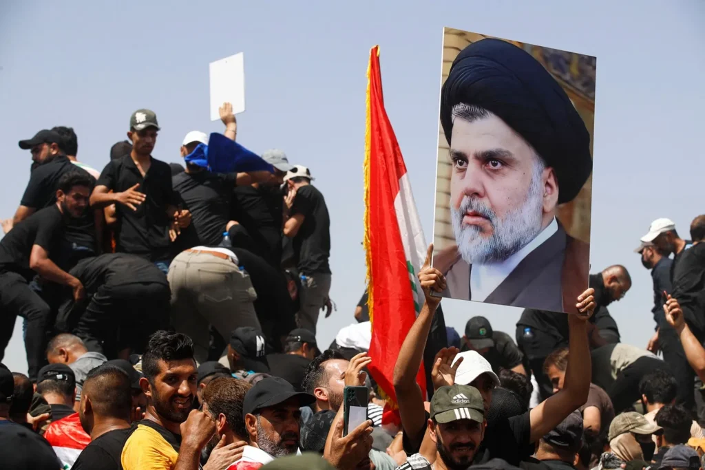 مقتدى الصدر يعلن دمج ميليشياته في الدولة العراقية A large crowd of supporters gathered during a public demonstration in Iraq, with one man holding up a large printed portrait of Muqtada al Sadr.