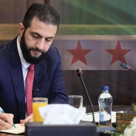 A close-up view of Syrian leader Ahmad al-Sharaa in a dark suit and red tie looking down while writing at a conference table with a microphone and a superimposed Syrian flag with two red stars, representing the administrative continuity of recent government appointments.