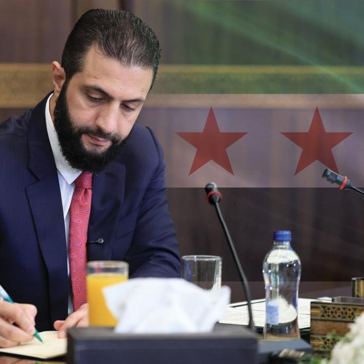 Syria’s New Government Appointments: More Continuity Than Change A close-up view of Syrian leader Ahmad al-Sharaa in a dark suit and red tie looking down while writing at a conference table with a microphone and a superimposed Syrian flag with two red stars, representing the administrative continuity of recent government appointments.