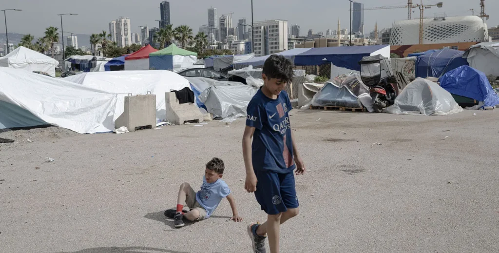 A Geographic and Social Reconfiguration in Lebanon Two young boys outdoors in an makeshift encampment area filled with tents and plastic tarps against a city skyline backdrop, visually documenting the displacement resulting from the Israel-Hezbollah conflict.