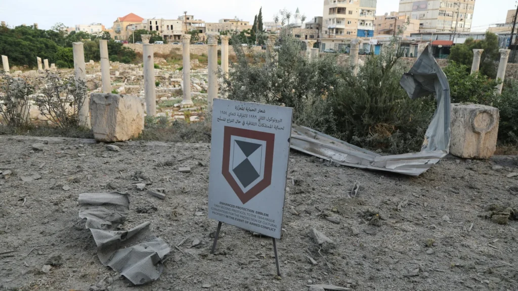 An 'Enhanced Protection Emblem' cultural property shield sign stands surrounded by debris and twisted metal near ancient Roman columns, demonstrating the severe destruction threatening Iran and Lebanon's ancient heritage.