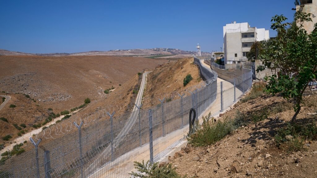 A heavily fortified border fence lined with razor wire stretches across a rugged desert landscape with a military watchtower and white outpost building in the distance, framing the tense security environment surrounding the war in Lebanon.