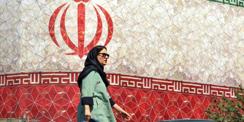 A woman wearing a headscarf and sunglasses walks past a large outdoor wall patterned with the national symbol and flag of Iran, providing a visual context for diplomatic talks regarding the memorandum of understanding.