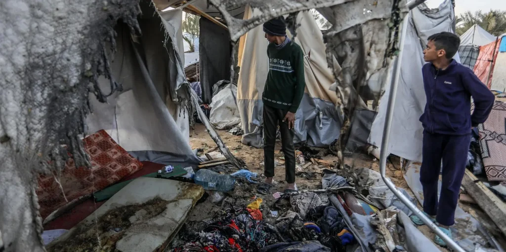 Two youth stand amidst the charred remains and scattered debris of destroyed tents within a makeshift camp in Gaza, surveying the severe devastation and illustrating the critical humanitarian conditions during this unstable phase of ceasefire.