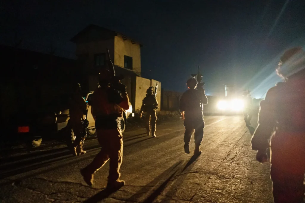 Armed soldiers in tactical gear moving along a dark road at night near a building, illuminated by bright vehicle headlights, representing the historical events tied to the Afghanistan reckoning.