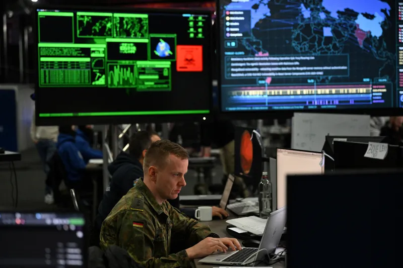 The Coming Quantum National Security Crisis A military officer in camouflage uniform sits at a workstation typing on a laptop in a dimly lit operations center with large cyberdefense monitoring screens in the background, illustrating the advanced environments central to preparing for a Quantum National Security crisis.