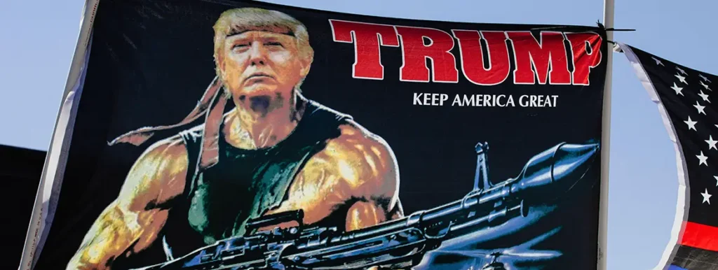 Peace Through Strength – How is that Working Out? A large black campaign banner depicting Donald Trump's face superimposed onto a muscular body holding a rocket launcher, featuring the text "TRUMP KEEP AMERICA GREAT" at a rally, symbolizing the political rhetoric surrounding the concept of peace through strength.