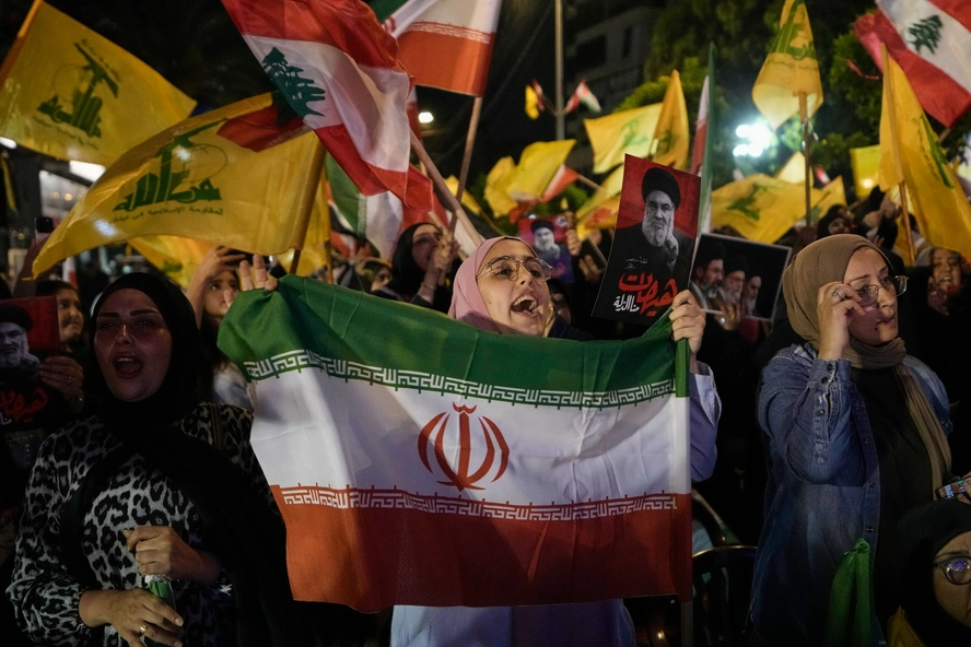 استراتيجية دبلوماسية الصواريخ لدى ترامب تنقلب عليه A crowd of supporters gathers at night holding yellow Hezbollah flags and Lebanese flags, with a woman in the center holding the official flag of the Islamic Republic of Iran, capturing the regional alignment reacting against Trump’s missile diplomacy.