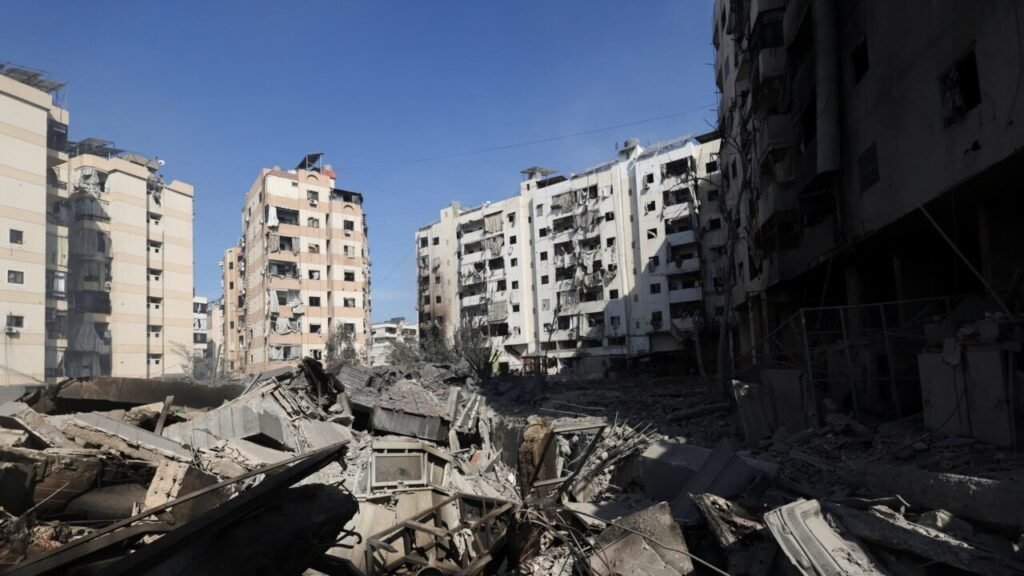 A wide shot of severe urban destruction with a massive pile of concrete rubble, exposed rebar, and debris in the foreground, flanked by heavily damaged multi-story residential buildings against a clear blue sky, illustrating the physical devastation along Iran's Battlefront in the Middle East.