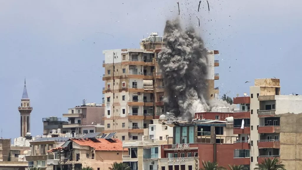 Dense gray smoke and debris exploding upward from a multi-story residential building following an airstrike in a coastal city, illustrating the destruction that highlights the urgent need for a meaningful Israel-Lebanon ceasefire.