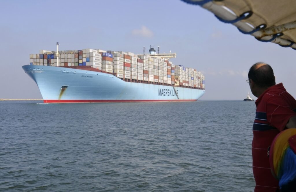 ترافیک به تدریج به کانال سوئز مصر بازمیگردد با وجود تهدید حوثیها A wide-angle view of a massive light blue Maersk container ship loaded with colorful cargo containers sailing on calm water, watched by a man in a red polo shirt from a vessel in the foreground, illustrating international maritime shipping routes returning to the Suez Canal.