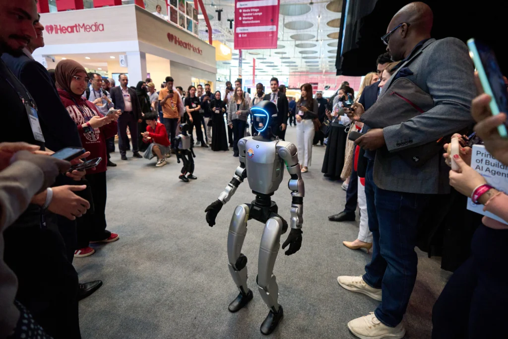 The new non-alignment: How the Middle East is carving out room to maneuver in AI A sleek bipedal humanoid robot with a glowing blue visor stands in the center of a crowded technology exhibition hall while attendees observe and film it with smartphones, showcasing regional technological advancements and the strategic maneuver in AI.