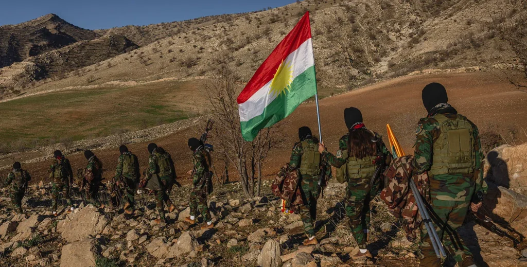 A line of masked fighters in military camouflage marching across a rocky mountain slope while holding a large Kurdish flag, representing armed combatants belonging to Kurdish Rebel Groups.