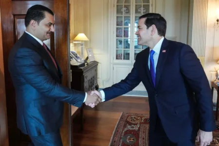 U.S. Secretary of State Marco Rubio smiles while shaking hands with Libyan National Army Deputy Commander Saddam Haftar inside a formal office setting, representing the high-level diplomatic meetings connected to an elite bargain.