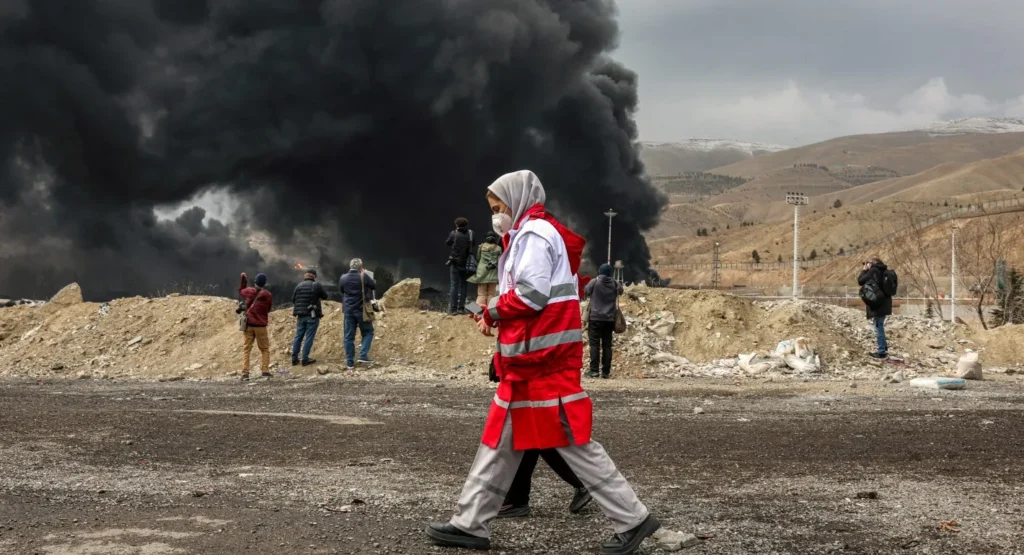 An Iranian Red Crescent worker walking past photographers near thick black smoke billowing from the targeted Shahran oil refinery near Tehran, illustrating damage to critical Energy Infrastructure.