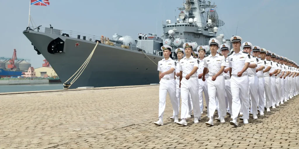 Thinking twice about that US-China superwar A long line of naval officers in crisp white uniforms march in unison along a paved dock next to a large grey warship docked at a port, symbolically highlighting the geopolitical tensions and military readiness surrounding a potential US-China superwar.