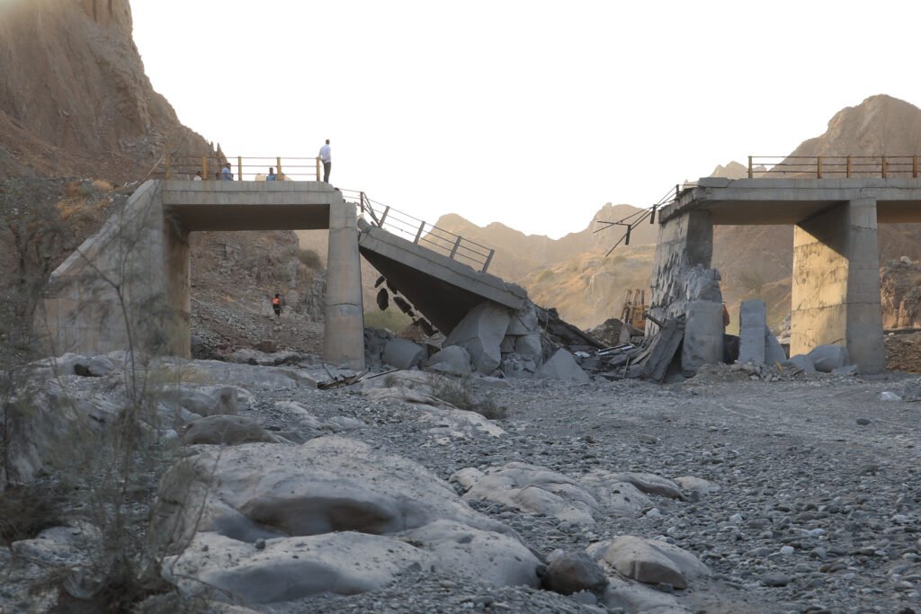 معضلة ترامب مع إيران: العودة إلى الحرب أم البحث عن مخرج People standing on the edge of a severely collapsed concrete bridge following a military strike in a mountainous region, illustrating consequences surrounding the Trump Iran Dilemma.