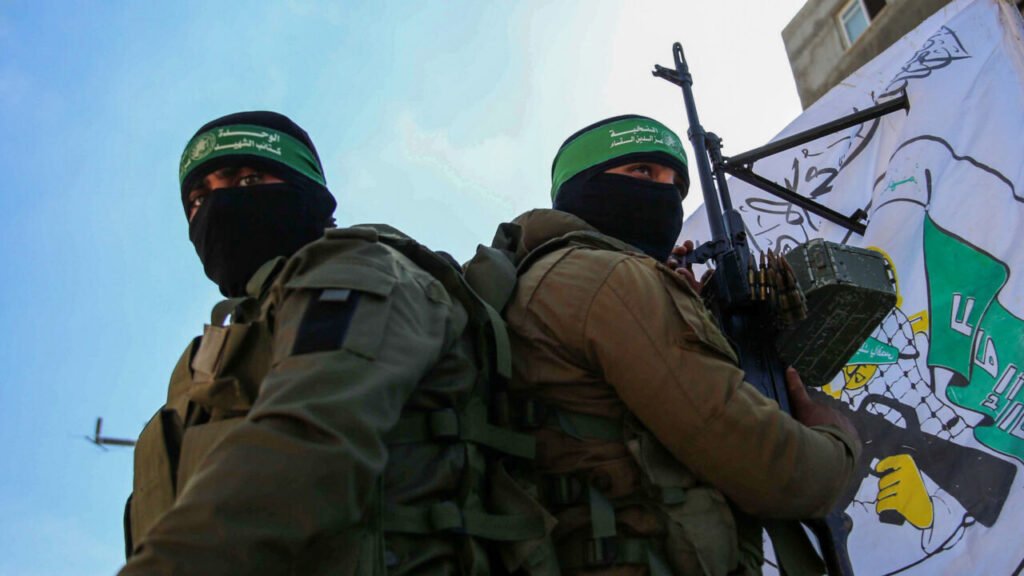 هل تستطيع إسرائيل تفكيك حماس؟ Two masked militants in tactical gear with green headbands, standing in front of a flag, representing the armed groups central to the question of whether Israel can Dismantle Hamas.