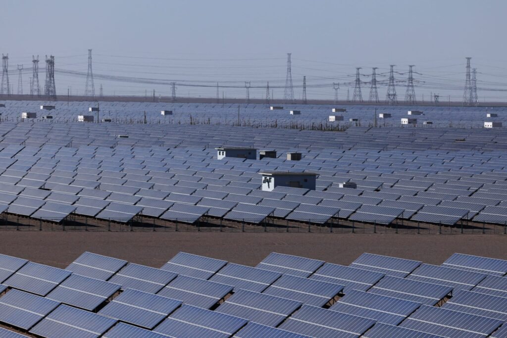 Why China Restrains Clean-Tech Edge in Hormuz Crisis Vast arrays of solar photovoltaic panels stretching across a desert landscape beneath power transmission towers in China, representing renewable energy infrastructure and Clean-Tech.