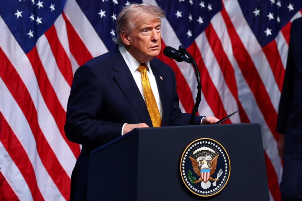 فروپاشی توافق ایران، ترامپ را با گزینههای محدود مواجه میکند A medium close-up shot of Donald Trump wearing a dark suit, white shirt, and yellow tie, speaking at a dark blue podium featuring the US presidential seal, with multiple American flags lined up in the background, capturing foreign policy challenges related to the Iran Deal Collapse.