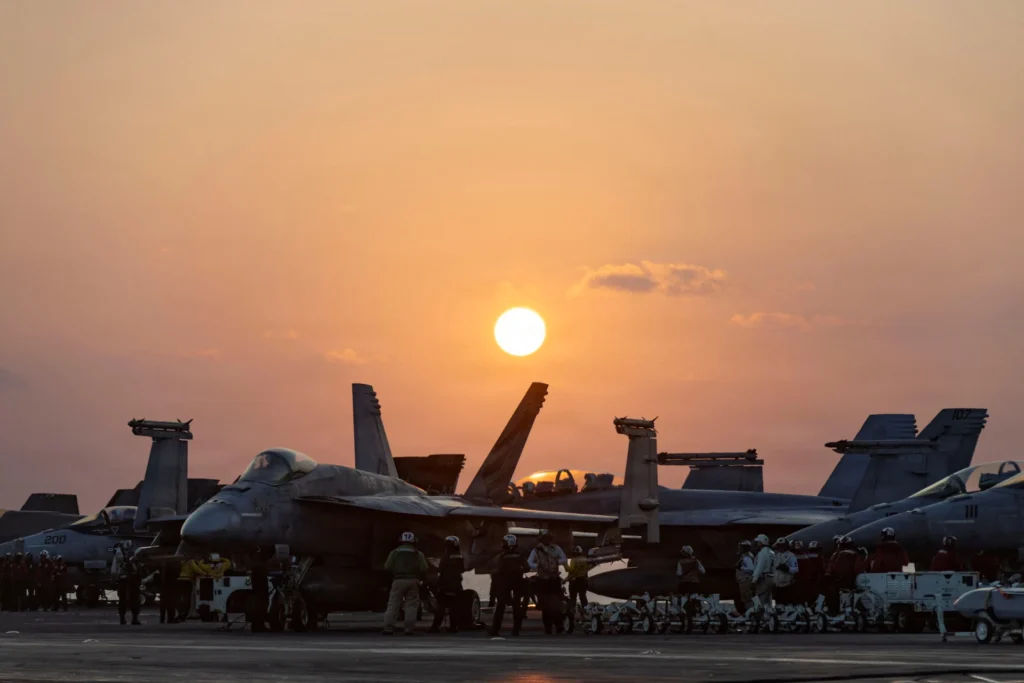 How Will the Iran War End? Trump’s Options U.S. Navy sailors preparing fighter jets on an aircraft carrier flight deck at sunset, representing naval strike operations tied to how the Iran War End.