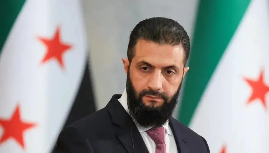 إزالة سوريا من قائمة الإرهاب لا تعني منحها تصريحًا مجانيًا A close-up portrait of Ahmed al-Sharaa wearing a dark suit and a red patterned tie with a serious expression, positioned between flags featuring red stars in the background, framing international debate over Syria's Terrorism.