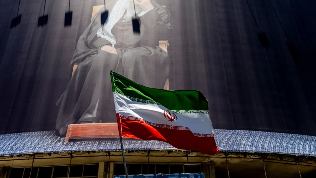The official flag of the Islamic Republic of Iran waves in the foreground below a large, towering monochrome banner depicting a seated religious leader, capturing the political environment surrounding discussions on why for Tehran, negotiation is war.