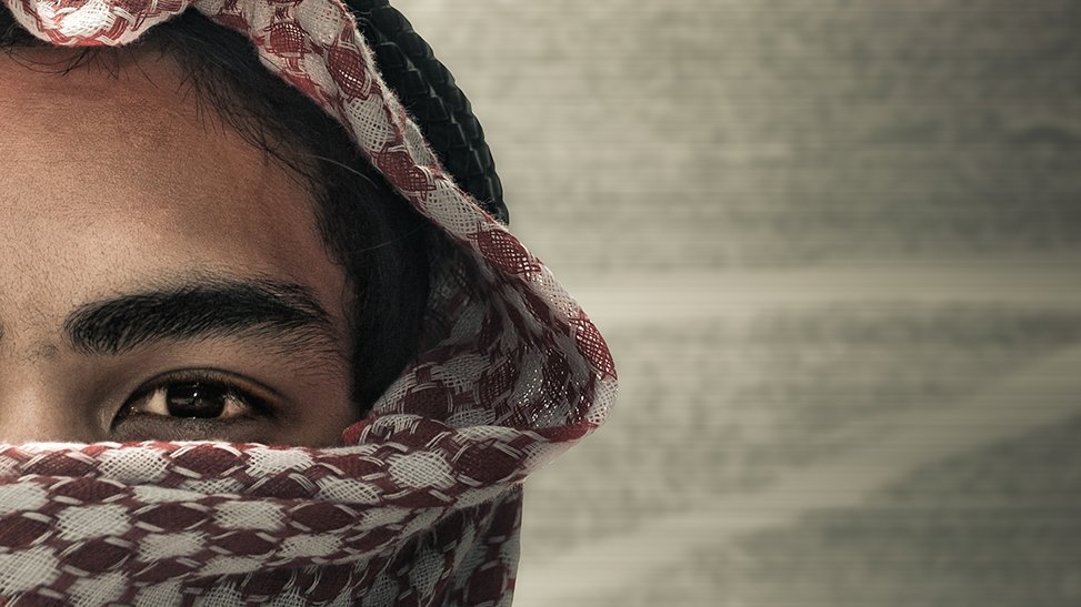 الشكل الجديد للجهاد العالمي A dramatic close-up cropped portrait of a young person whose lower face is covered by a patterned red and white keffiyeh or headscarf, leaving only one eye and eyebrow visible against a blurred rustic background, capturing thematic concepts linked to Global Jihadism.