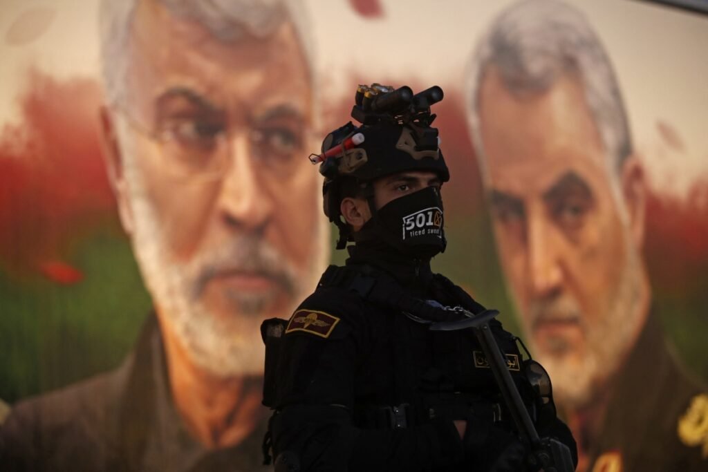 A soldier in black tactical gear, helmet, and a face mask stands in the foreground, with large blurred portraits of Abu Mahdi al-Muhandis and Qasem Soleimani visible on a billboard in the background, illustrating regional dynamics surrounding the Iranian crescent.